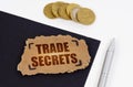 On a black and white surface lies a pen, coins and a cardboard sign with the inscription - TRADE SECRETS