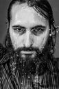 Black and white very close-up portrait of a young bearded man with soggy face and intense gaze with streams of water running down