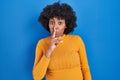 Black woman with curly hair standing over blue background asking to be quiet with finger on lips Royalty Free Stock Photo