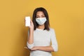 Black woman in medical face mask holding smart phone with empty display on vivid yellow background