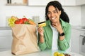 Black woman showcases credit card for grocery shopping in kitchen