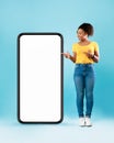 Black woman standing next to huge smartphone, pointing at empty screen, advertising new mobile app, mockup Royalty Free Stock Photo
