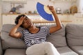 Black woman suffering from heatstroke flat without air-conditioner waving fan lying on couch at home