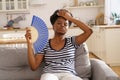 Black woman suffering from heatstroke flat without air-conditioner waving fan lying on couch at home