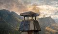 Black wood smoke coming out of the chimney of a roof of a rural town house in the middle of some mountains with cloudy sky at Royalty Free Stock Photo