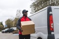 Black young adult delivery guy wearing work uniform and black cap smiling walking by white van holding carboard box Royalty Free Stock Photo