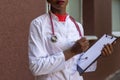 Black young woman doctor, in a white coat, with a phonendoscope, holds a folder for medical papers in a rue, against the Royalty Free Stock Photo