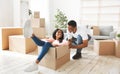 Black young woman riding carton box, having fun with her boyfriend in new apartment, panorama