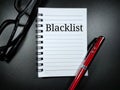 Blacklist in black word on notebook with pen and glasses on black background.
