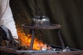 Blacksmith heating up metal in red hot coals while tea pot boiling water on top. Craftsman hand forging metal, red sparks are