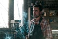 Blacksmith in leather apron smokes a cigarette at pause in work on the background of workspace