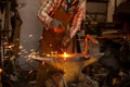 The blacksmith manually forging the molten metal on the anvil in smithy with spark fireworks.