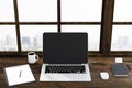 Blank black laptop screen, coffee mug, and glasses on wooden table with city view window