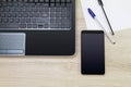 Blank black screen smartphone beside laptop with paper and pens on wooden desk Royalty Free Stock Photo