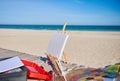 White blank canvas on a wooden table easel next to paints and brushes on a wooden deck chair on the beach against the beautiful Royalty Free Stock Photo