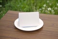 Blank folded card on a classic empty white plate on the  outdoor wooden table and green grass background behind, clean template Royalty Free Stock Photo