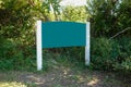 Blank green wooden sign on white posts with large bushes in the background