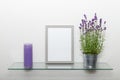 Blank grey wooden photo frame, lavender flowers in zinÃÂ pot, violet vintage candle on glass shelf against light wall Royalty Free Stock Photo