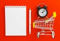 Blank notepad and pen, alarm clock and mini supermarket cart on red background. Online shopping, list, time saving concept. Flat Royalty Free Stock Photo