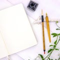 Blank page of sketchbook with calligraphy tools and green foliage. Notebook top view photo on white background