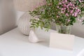 Blank paper card, bunch of flowers in vase, bird, table lamp on white table. Happy Mothers day greeting card mockup in