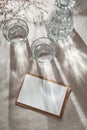 Blank paper card with copy space, glasses and bottle with water or alcohol on neutral beige table, aesthetic sunlight shadows,