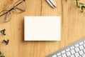 Blank paper card mockup on wooden office desk table with female glasses, office supplies, computer keyboard. Top view, flat lay,