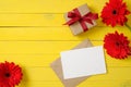 Blank paper card, vintage envelope, red daisy flowers, gift box on yellow wooden background. Women desk. Congratulation or Royalty Free Stock Photo