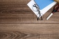 A blank sheet of paper, a notebook, pens, glasses, scissors on a wooden table top, space for text