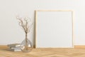 Blank square poster frame mock up standing on light herringbone parquet floor next to white wall with vase and books.