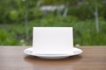 Blank standing bifold card on a classic empty white plate on the outdoor wooden table and green garden behind, clean template for Royalty Free Stock Photo