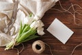 Blank wedding invitation card and bunch of spring flowers on wooden table. White tulips, textile rag, twine, romantic wedding gree