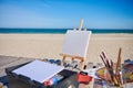 Blank white canvas on a wooden table easel next to paints and brushes on a wooden deck chair on the beach against the background Royalty Free Stock Photo