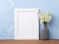 Blank white frame, flower in vaze on brown wooden table against pastel blue concrete wall with copy space. Mock up. Royalty Free Stock Photo