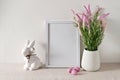 Blank white picture frame mockup, flowers in vase, rabbit figurine, pink candy eggs on neutral beige table background Royalty Free Stock Photo