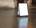 Blank white sandwich sign outside in daytime on dark wet sidewalk near a building.