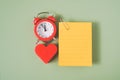 Blank yellow note paper with paper clip, red heart shape and analog alarm clock on green background Royalty Free Stock Photo