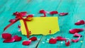 Blank yellow place card with red ribbon and rose petals on antique teal blue distressed wood table Royalty Free Stock Photo