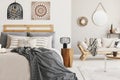 Blanket on bed under lamp and wooden stool in boho bedroom interior with poster and sofa