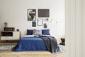 Blanket on blue bed with pillows in retro bedroom interior with radio on cabinet and posters. Real photo