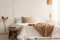 Blanket on white bed in natural bedroom interior with plants and wooden stool