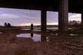 A bleak, moody, winter edit of a figure standing next to a lake, looking out under a motorway bridge at sunset