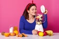 Blender with berries and fruit on table. Female makes tasty smoothie and likes its smell. Charming girl wears blue sweater and