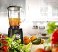 A blender on the table with some fruit and vegetables with blurred kitchen background.