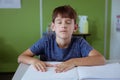 Blind caucasian schoolboy sitting in classroom with eyes closed reading braille book with fingers