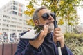 Blind middle-aged man with gray hair and beard wearing sunglasses holding cane and phone using text-to-speech function