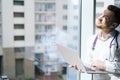 Blinking from sun male doctor in white uniform with stethoscope using laptop holding with one hand, standing near window