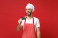 Blinking young bearded male chef cook or baker man in striped apron white t-shirt toque chefs hat isolated on red wall Royalty Free Stock Photo