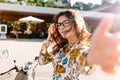 Blissful brunette girl in bright shirt making selfie in front of restaurant in summer day. Portrait of charming young Royalty Free Stock Photo