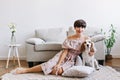 Blissful brunette girl in cute outfit sits on carpet in front of gray sofa with her puppy. Indoor portrait of smiling Royalty Free Stock Photo